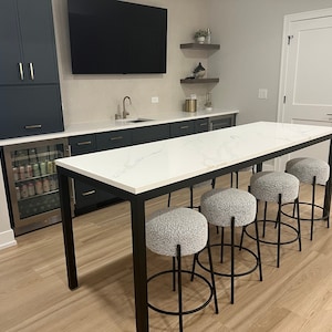 May include: A white countertop bar table with black metal legs and five grey upholstered bar stools. The table is in a basement with a dark blue cabinet and a mini fridge.