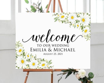 Op de afbeelding: Een wit welkomsbord voor een bruiloft met een bloemenontwerp met witte madeliefjes en groene bladeren. Het bord staat "Welkom op onze bruiloft Emilia & Michael 25 augustus 2021".