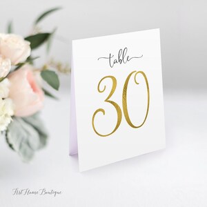 Könnte beinhalten: Weiße Tischnummerkarte mit goldfarbener Folienbeschriftung. Die Karte lautet "Tisch 30".