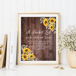 May include: A rustic wooden sign with a sunflower and baby's breath design. The sign reads "A Bucket List for the Bride and Groom! Leave a note, idea, or plan but more importantly an adventure we promise to complete it in a lifetime!"
