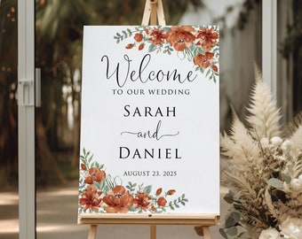 May include: A white rectangular wedding welcome sign on a wooden easel featuring watercolor rust-colored flowers and green foliage. The text reads Welcome to our wedding, Sarah and Daniel, and the date August 23, 2025, in black serif and script fonts.