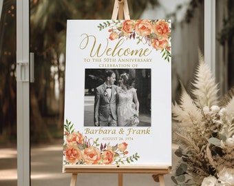 May include: A white welcome sign on an easel, featuring a photo of a couple and floral accents. The sign reads "Welcome to the 50th Anniversary Celebration of Barbara & Frank August 24, 1974". The floral design includes orange and peach-colored roses.