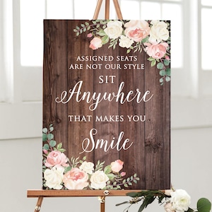 May include: A wooden sign on an easel with the text "ASSIGNED SEATS ARE NOT OUR STYLE SIT ANYWHERE THAT MAKES YOU SMILE." The sign is decorated with floral arrangements of pink and white roses and greenery.