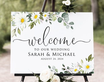 Op de afbeelding: Een wit trouwbord op een zwart schildersezel, versierd met bloemstukken. Het bord toont het woord "welcome" in elegant schrift, samen met de namen Sarah & Michael en de datum 24 augustus 2024.