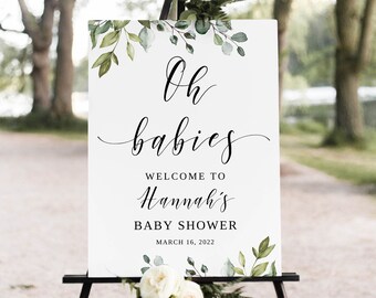 May include: A white sign with green foliage and the text "Oh babies" in black cursive font. The sign also says "Welcome to Hannah's Baby Shower" and "March 16, 2022". The sign is on a black easel.