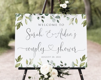 May include: A white welcome sign with green foliage border, featuring the text "Welcome to Sarah & Aiden's couples shower August 20, 2022".