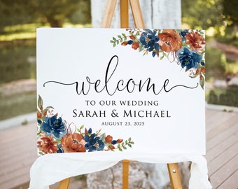 May include: A white wedding welcome sign on a wooden easel. The sign has floral arrangements in blue, rust, and green. The text reads "welcome to our wedding" followed by the names "Sarah & Michael" and the date "August 23, 2025".