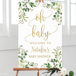 May include: A white welcome sign with gold lettering that reads "Oh baby, Welcome to Natalia's Baby Shower". The sign is decorated with green watercolor leaves and branches.