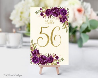 May include: Table number 56 with a gold font on a cream background. The number is surrounded by a wreath of purple and green watercolor flowers.