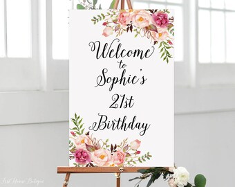 May include: A white rectangular birthday sign featuring floral illustrations in shades of pink, peach, and green at the top and bottom corners. The black text reads Welcome to Sophie's 21st Birthday in a decorative script font on a smooth surface.