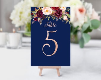 May include: A navy blue table number 5 card with floral accents. The card features a rose gold number 5 and the word "Table" in a script font. The top of the card is decorated with burgundy, cream, and green flowers.