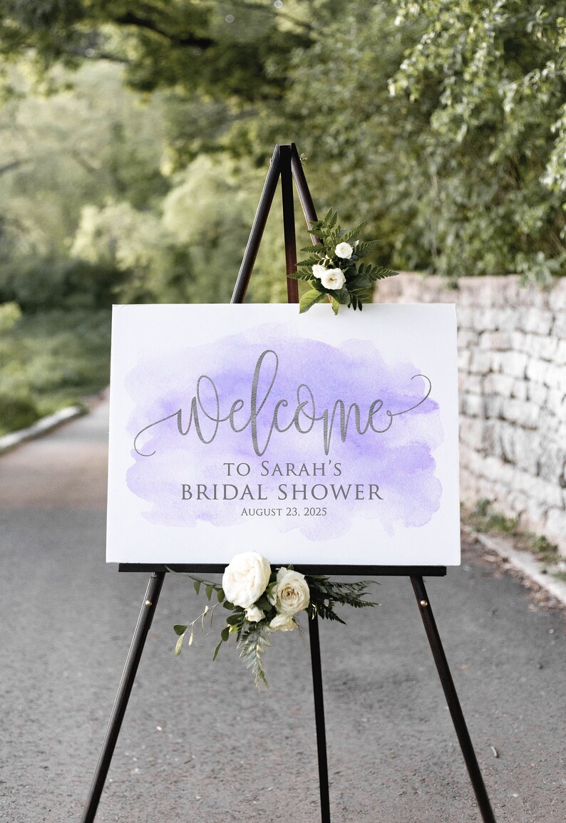 May include: A white sign with a purple watercolor background on an easel. The sign reads 'Welcome to Sarah's Bridal Shower August 23, 2025'.