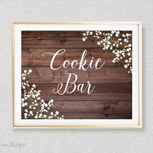 May include: A rustic wooden sign with the text "Cookie Bar" in white cursive font. The sign is decorated with white baby's breath flowers in the corners.