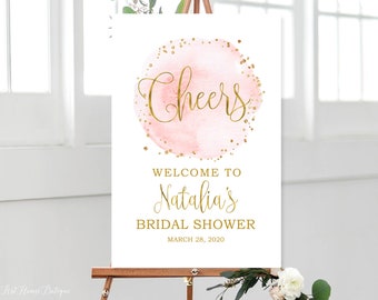 May include: A white welcome sign with gold lettering for a bridal shower. The sign features a pink watercolor circle with gold glitter and the word "Cheers". The text reads "Welcome to Natalia's Bridal Shower March 28, 2020".