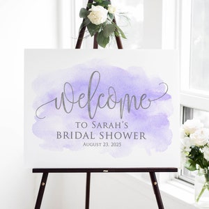 May include: A white sign with a purple watercolor background and silver lettering. The sign reads 'Welcome to Sarah's Bridal Shower August 23, 2025.' The sign is displayed on a wooden easel.