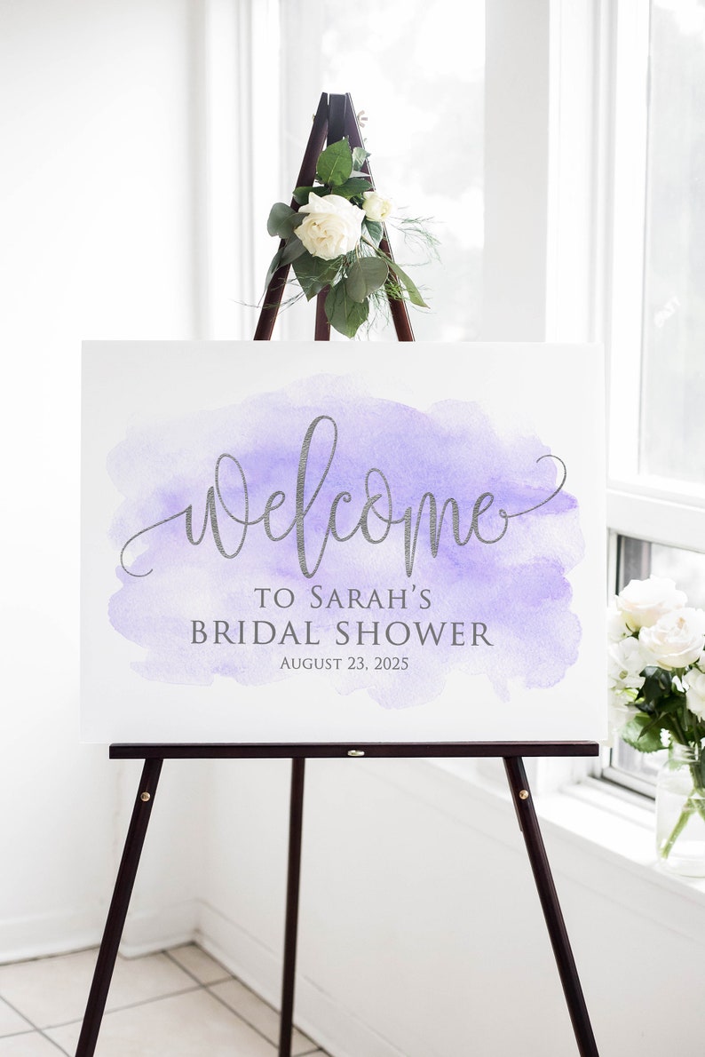 May include: A white sign with a purple watercolor background and silver lettering. The sign reads 'Welcome to Sarah's Bridal Shower August 23, 2025.' The sign is displayed on a wooden easel.
