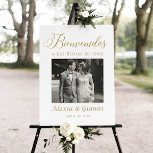 Op de afbeelding: Een wit welkomsbord met gouden letters met de tekst "Bienvenidos a las bodas de oro" met een zwart-witfoto van een stel en de tekst "Alexia & Gianni Abril 20, 2024".