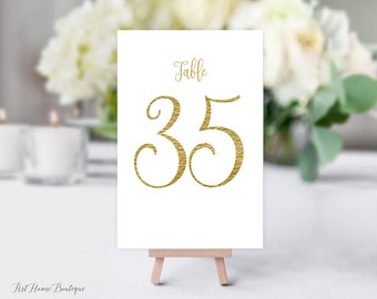 May include: Gold table number card with the number 35 in a decorative script font. The card is on a small wooden easel.