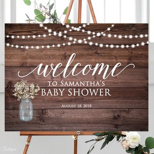 May include: Rustic wooden welcome sign with white text that reads "Welcome to Samantha's Baby Shower August 18, 2018". The sign features a mason jar with white flowers and string lights.