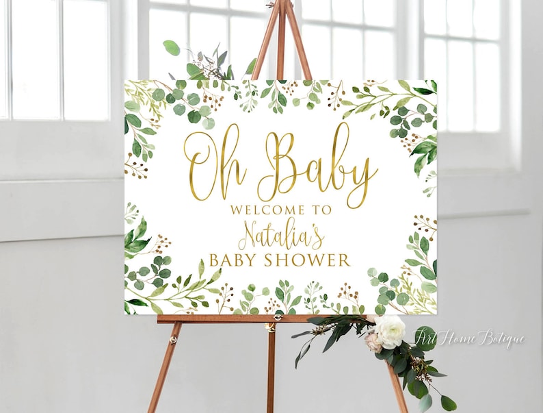 May include: A white welcome sign with gold lettering that reads "Oh Baby Welcome to Natalia's Baby Shower". The sign is decorated with green watercolour leaves and branches.