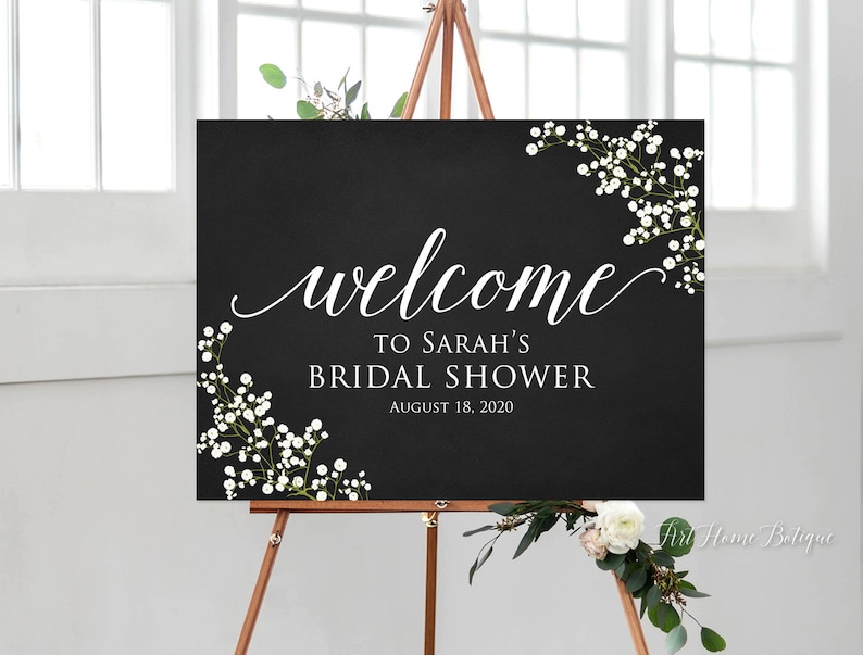 May include: A black sign with white script that reads "welcome to Sarah's Bridal Shower August 18, 2020." The sign is decorated with white flowers and greenery in the corners. The sign is displayed on a wooden easel.