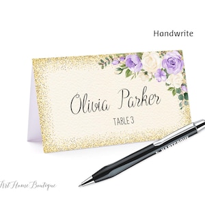May include: A place card with gold glitter trim, featuring the name "Olivia Parker" and "Table 3". The card is decorated with purple and white floral accents. A black pen rests on the surface.
