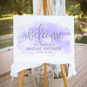 May include: A white welcome sign with a purple watercolor background. The sign reads 'Welcome to Sarah's Bridal Shower August 23, 2025'.
