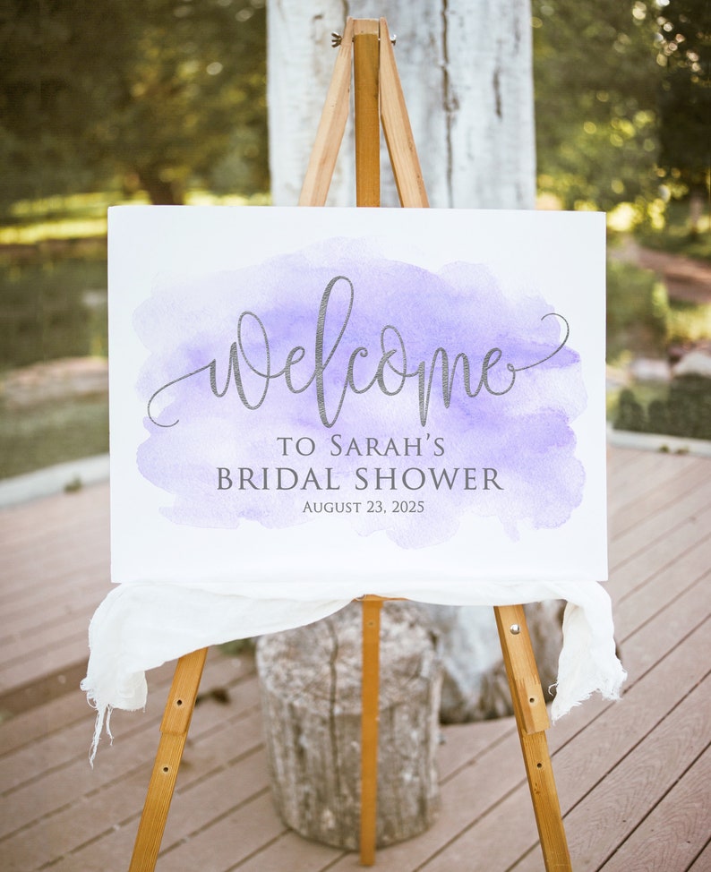 May include: A white welcome sign with a purple watercolor background. The sign reads 'Welcome to Sarah's Bridal Shower August 23, 2025'.
