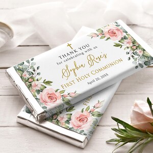 May include: Two chocolate bars with silver foil wrapping. The white wrappers feature floral designs with pink roses and green leaves. Gold text reads "THANK YOU," "Sophia Rose's," and "FIRST HOLY COMMUNION."