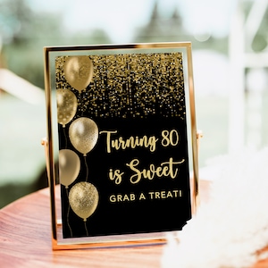 May include: A black and gold sign with gold balloons and glitter. The sign reads "Turning 80 is Sweet Grab a Treat!"