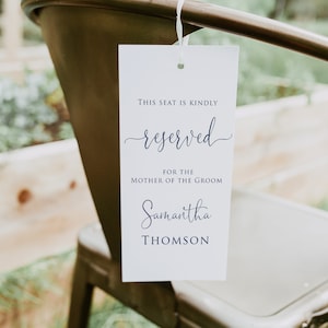 May include: White card with blue text that reads "This seat is kindly reserved for the mother of the groom Samantha Thomson". The card is hanging from a metal chair.
