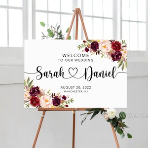 K&ouml;nnte beinhalten: Ein wei&szlig;es Hochzeitsschild mit burgunderroten und blushfarbenen floralen Akzenten. Das Schild lautet "Willkommen zu unserer Hochzeit Sarah Daniel 20. August 2022 Manchester, NJ".