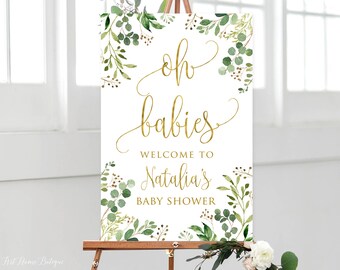 May include: A white welcome sign with gold lettering that reads "Oh babies, Welcome to Natalia's Baby Shower". The sign is decorated with green watercolor leaves and branches.