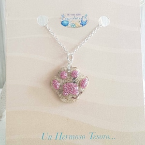 May include: A silver chain necklace with a pink glitter paw print pendant made of sand and resin. The pendant is on a white card with the text "Un Hermoso Tesoro..." and the logo "LagunArteFR".