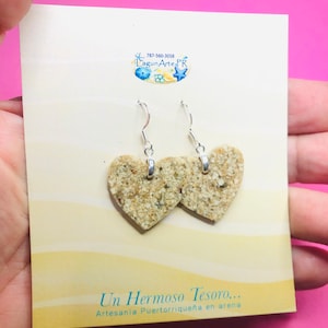 May include: A pair of heart-shaped earrings made with sand from Puerto Rico. The earrings are silver and have a natural, sandy texture. The card reads "Un Hermoso Tesoro... Artesania Puertorriqueña en arena".