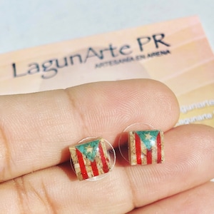 May include: A pair of small square earrings with the Puerto Rican flag embedded in the center. The earrings are made of resin and have a clear backing.