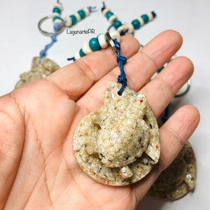 May include: A close-up of a sea turtle keychain made of sand and resin. The keychain is attached to a blue cord and a silver key ring. The turtle is surrounded by white and blue beads.