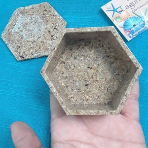 Puede incluir: Una caja hexagonal hecha de materiales naturales, como conchas marinas y arena. La caja tiene una tapa y está abierta, revelando el interior. La caja está sentada en la mano de una persona.