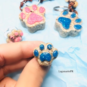 May include: Three resin paw print keychains with glitter embedded in the paw prints. The keychains are in pink, blue, and blue. The blue paw print keychain is being held in a hand.