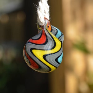 Op de afbeelding: Een kleurrijke glazen hanger met een draaiende patroon van rood, geel, zwart en blauw. De hanger is bevestigd aan een wit koord.