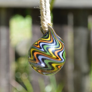 Puede incluir: Un colgante de vidrio colorido con un patrón de remolino de negro, verde, amarillo y naranja. El colgante está colgado de una cuerda blanca.