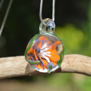 Può includere: Una collana con un ciondolo di vetro su una catena d'argento. Il ciondolo è una sfera di vetro trasparente con un motivo floreale colorato all'interno. Il fiore è arancione, blu e bianco.