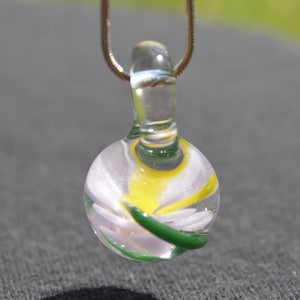Può includere: Una collana con un ciondolo di vetro, una balla di vetro trasparente e una sfera rotonda di vetro trasparente che contiene un vortice di vetro bianco, giallo e verde.
