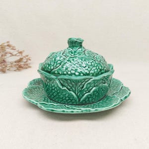 Peut inclure: Un bol en céramique verte avec couvercle en forme de chou, présentant un design extérieur texturé et feuillu. Le bol repose sur une soucoupe en céramique verte assortie qui imite l'apparence de grandes feuilles de chou superposées et brillantes.