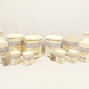 Può includere: Una collezione di barattoli e bottiglie da cucina vintage in ceramica color crema. Ogni pezzo presenta un design floreale con accenti blu e rosa e finiture dorate. Alcuni pezzi hanno etichette tedesche.