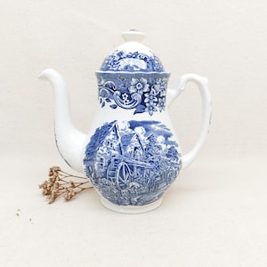 Może przedstawiać: Biały ceramiczny imbryk z niebieskim kwiatowym wzorem na pokrywce i uchwycie. Korpus przedstawia niebiesko-białą scenę pasterską. Imbryk ma długi dzióbek i zakrzywiony uchwyt.