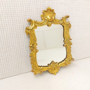 Può includere: Uno specchio da parete rettangolare con una cornice color oro metallizzato decorata con intricati motivi a volute e intagli a forma di foglia. La cornice presenta un prominente fregio decorativo al centro in alto e un dettaglio simile in basso.