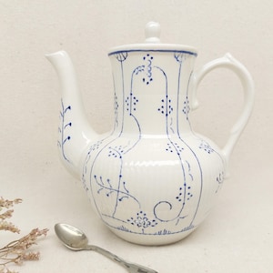 Può includere: Teiera in ceramica bianca con coperchio, beccuccio e manico. La teiera è decorata con motivi floreali blu. Un cucchiaio d'argento è appoggiato vicino alla teiera. La teiera è un oggetto decorativo.