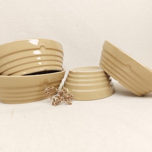 Może przedstawiać: Zestaw trzech beżowych misek ceramicznych z błyszczącym wykończeniem i poziomymi żebrowanymi detalami na zewnętrznych ściankach. Miski są ułożone w grupę, dwie ustawione jedna na drugiej po lewej stronie i jedna pionowo po prawej stronie.