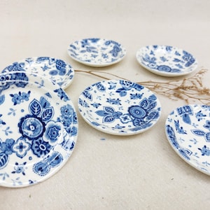 Peut inclure: Six petites assiettes en céramique blanche avec un motif floral bleu. Les assiettes ont un fond blanc avec des fleurs et des feuilles bleues.